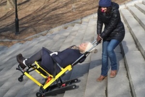 A person in a coat assists another person seated in a yellow mobile stairlift on outdoor steps.