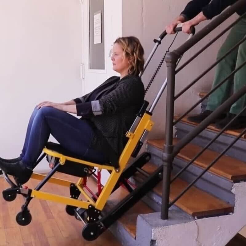 A person uses a yellow, mobile stair climbing wheelchair to descend stairs, assisted by another person controlling the chair from behind.